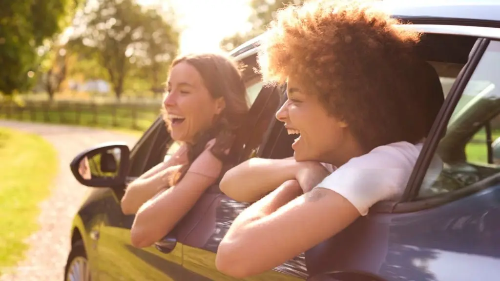 Two friends laughing as they lean out of a car window on a sunny drive
